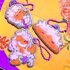 May include: Three colourful keychains featuring cartoon characters with fluffy white hair. The characters are all smiling and have different expressions. The keychains are made of plastic and have a rainbow-coloured border.