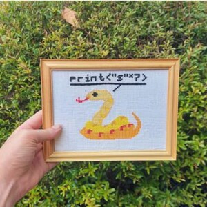 Op de afbeelding: Een ingelijst kruissteekontwerp met een gele slang met rode accenten. De tekst "print("s")" is boven de slang geborduurd.
