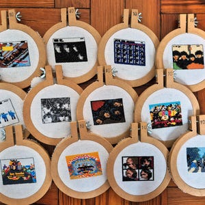 Könnte beinhalten: Zehn Kreuzstich-Kunstwerke, jeweils in einem hölzernen Stickrahmen montiert. Die Designs zeigen pixelige Bilder von Albumcovern in verschiedenen Farben und Stilen. Die Rahmen sind auf einer Holzoberfläche angeordnet.