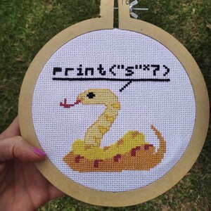 Op de afbeelding: Een witte kruissteek borduurring met een gele slang met een rode tong en de tekst "print('s' * 7)" in zwart garen.