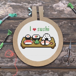 Puede incluir: Un aro de bordado blanco con un marco de madera marrón. El bordado representa un plato de sushi con el texto "I ♥ sushi" encima.