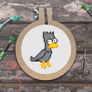 Puede incluir: Un aro de bordado de punto de cruz con un pájaro gris con patas amarillas y un pico amarillo. El pájaro tiene una cabeza puntiaguda y ojos grandes.