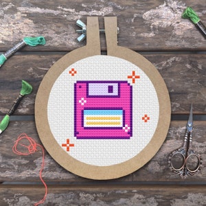 Puede incluir: Bordado de punto de cruz de un disquete retro en un aro de madera. El diseño presenta un disco rosa con rayas azules, amarillas y blancas. Hilo de bordar verde, tijeras y un fondo de madera completan la escena artesanal.