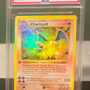 May include: A 1999 Pokemon Game Charizard Holo trading card graded GEM MT 10 by IPSA. The card features a yellow border, a holographic illustration of the orange dragon-like creature, 120 HP, and text detailing the Energy Burn power and Fire Spin attack.