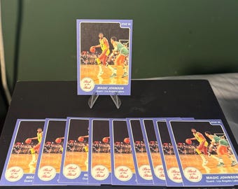 Könnte beinhalten: Eine Sammlung von Magic Johnson Basketballkarten. Die Karten zeigen ein Bild von Magic Johnson in einem gelb-goldenen Trikot beim Dribbeln eines Basketballs. Die Karten liegen auf einer schwarzen Oberfläche mit grünem Hintergrund.