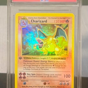 May include: A graded 1999 Pok&eacute;mon Game Charizard-Holo card. The card features a holographic image of Charizard, a flame Pok&eacute;mon, with a length of 5' 7" and a weight of 200 lbs. The card is encased in a protective case.