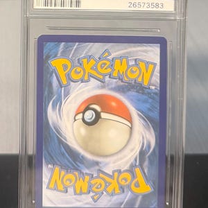 May include: A graded Pok&eacute;mon trading card in a clear plastic case. The card's back features the Pok&eacute;mon logo in yellow and a red and white Pok&eacute; Ball. The case has an IPSA label and a QR code.