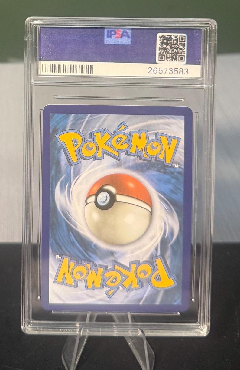 May include: A graded Pok&eacute;mon trading card in a clear plastic case. The card's back features the Pok&eacute;mon logo in yellow and a red and white Pok&eacute; Ball. The case has an IPSA label and a QR code.