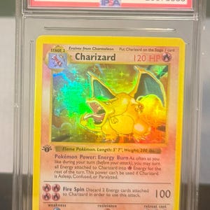 May include: A graded 1999 Pok&eacute;mon Game Charizard-Holo card. The card features a holographic image of Charizard, a flame Pok&eacute;mon, with a length of 5' 7" and a weight of 200 lbs. The card is encased in a protective case.