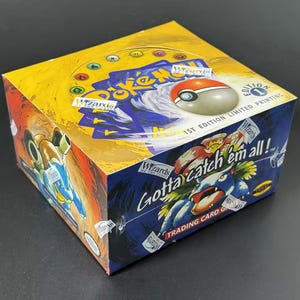 Puede incluir: Una caja sellada del juego de cartas coleccionables Pokémon. La caja es amarilla y azul con el texto "Gotta Catch 'Em All!" y "1st Edition Limited Printing". La caja presenta imágenes de personajes Pokémon y una Pokéball.