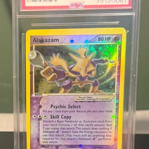 May include: A graded Alakazam-Holo Pokémon card from the 2006 Gold Star series. The card features a holographic image of Alakazam with a purple and yellow color scheme. The card is encased in a protective holder with a red label.