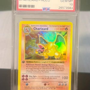 May include: A graded 1999 Pok&eacute;mon Game Charizard-Holo card. The card features a yellow Charizard illustration with a holographic background. The card is encased in a clear protective holder with the text "1999 POKEMON GAME CHARIZARD-HOLO" and "#4 GEM MT 10".