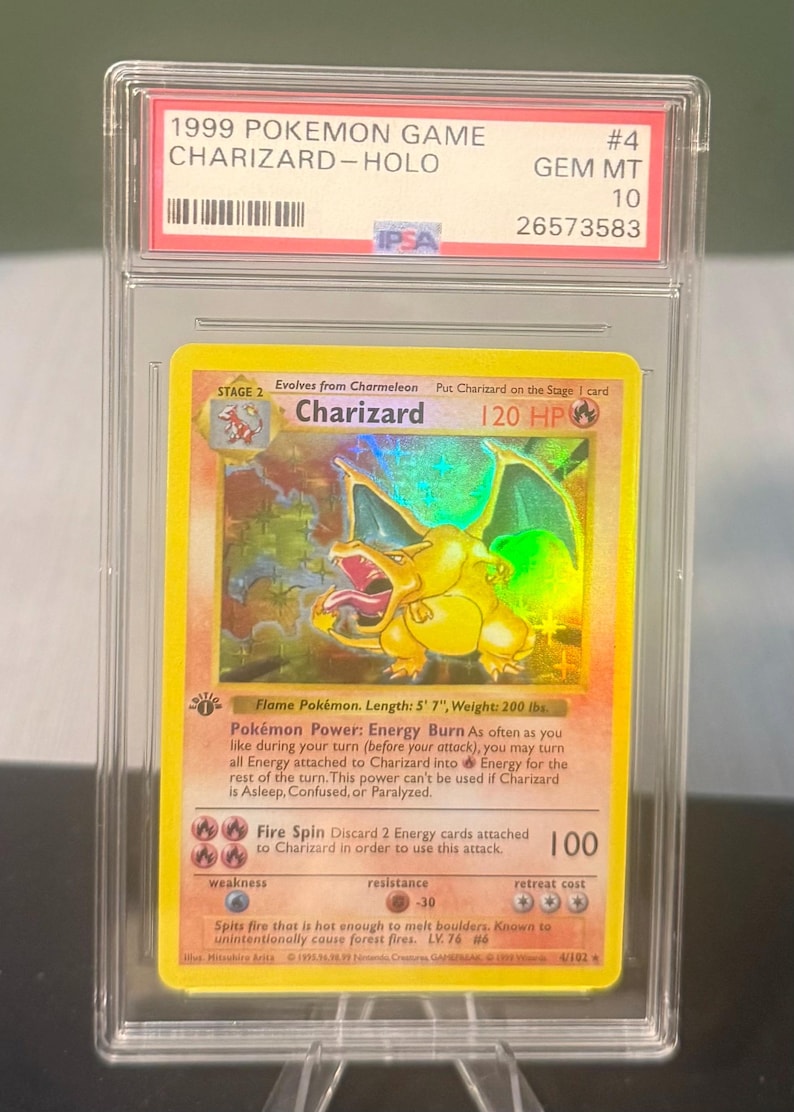 May include: A graded 1999 Pok&eacute;mon Game Charizard-Holo card. The card features a yellow Charizard illustration with a holographic background. The card is encased in a clear protective holder with the text "1999 POKEMON GAME CHARIZARD-HOLO" and "#4 GEM MT 10".