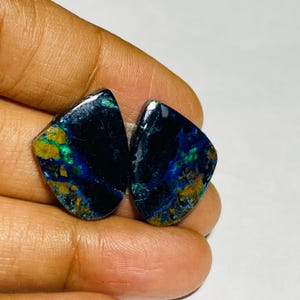 May include: Two polished gemstones with a dark blue base colour, accented with swirls of green and gold. The stones have a unique, irregular shape and a glossy finish. The gemstones are held in a hand.