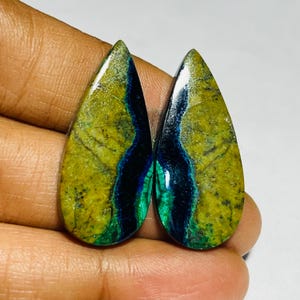 May include: Two polished, teardrop-shaped gemstones. The stones display a mix of olive green, dark blue, and green veins. The gemstones have a glossy finish. The overall effect is of natural stone with a unique pattern.