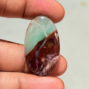 Peut inclure: Une pierre précieuse polie de forme ovale présentant une surface lisse avec des zones de couleurs distinctes. La partie supérieure est d'un vert menthe pâle, tandis que la section inférieure affiche des teintes brun-rouge et violet foncé.