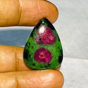 Może przedstawiać: Kształtny kamień szlachetny w kształcie łzy z zieloną podstawą i nakrapianymi wzorami. Widoczne są dwie głęboko czerwone inkluzje rubinu. Wypolerowana powierzchnia odbija światło, podkreślając naturalne piękno kamienia.