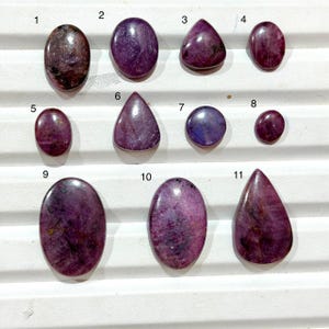 May include: Eleven polished cabochon gemstones in various shapes including oval, round, and teardrop. The stones display deep shades of purple, magenta, and dark red with natural inclusions and smooth, reflective surfaces, numbered one through eleven.