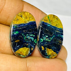 May include: Two oval azurite malachite gemstones. The stones display a striking mix of colours, including deep blues, greens, and yellows. The patterns resemble a landscape with a blue sky, green water, and yellow land.