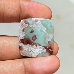 Peut inclure: Une pierre précieuse polie de forme carrée présentant un motif marbré de vert menthe, de blanc et de brun rougeâtre profond. La pierre affiche des bandes complexes, ondulées et superposées sur toute sa surface, avec deux inclusions brun foncé.