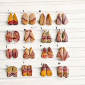 May include: A collection of sixteen pairs of polished, multi-colored jasper cabochons arranged in a grid. Each pair features unique natural patterns, including shades of yellow, pink, red, and tan, in various shapes like teardrops and ovals.