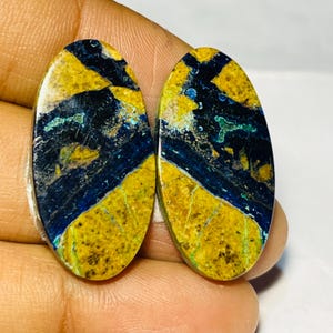 May include: Two oval gemstones with a striking pattern. The stones feature a mix of colours, including yellow, deep blue, and black, with veins of green. The polished surfaces showcase a natural, marbled effect, ideal for jewellery or crafting.
