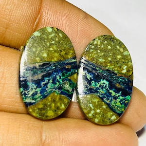May include: Two polished, oval gemstones. The stones display a mix of green, blue, and brown colours. The top section is speckled green, while the bottom has a mix of blue and green. The stones are held in a hand.