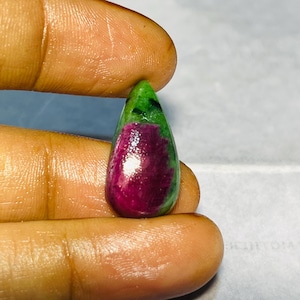 Può includere: Una gemma a forma di goccia con una vibrante combinazione di colori. La parte superiore è di un verde intenso, che sfuma in un magenta ricco e saturo nella metà inferiore. La superficie lucida riflette la luce, evidenziando la bellezza naturale della pietra.