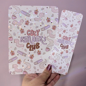 May include: A notebook and bookmark set with a repeating pattern of books, candles, and coffee cups. The words "Cozy Readers Club" are written in pink and brown. The items are held by a person.