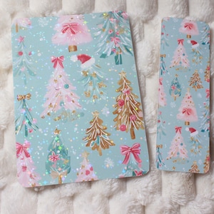 May include: Two patterned items featuring a light blue backdrop and a repeating design of Christmas trees in pink, gold, and green. The trees are decorated with bows, baubles, and Santa hats. One item is rectangular, the other is a smaller, elongated rectangle.
