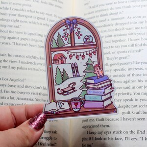 May include: A winter-themed sticker showing a window scene with a snowy landscape, trees, skis, and a gingerbread man on books. A mug of cocoa and an open book are also depicted. The sticker is held over an open book.