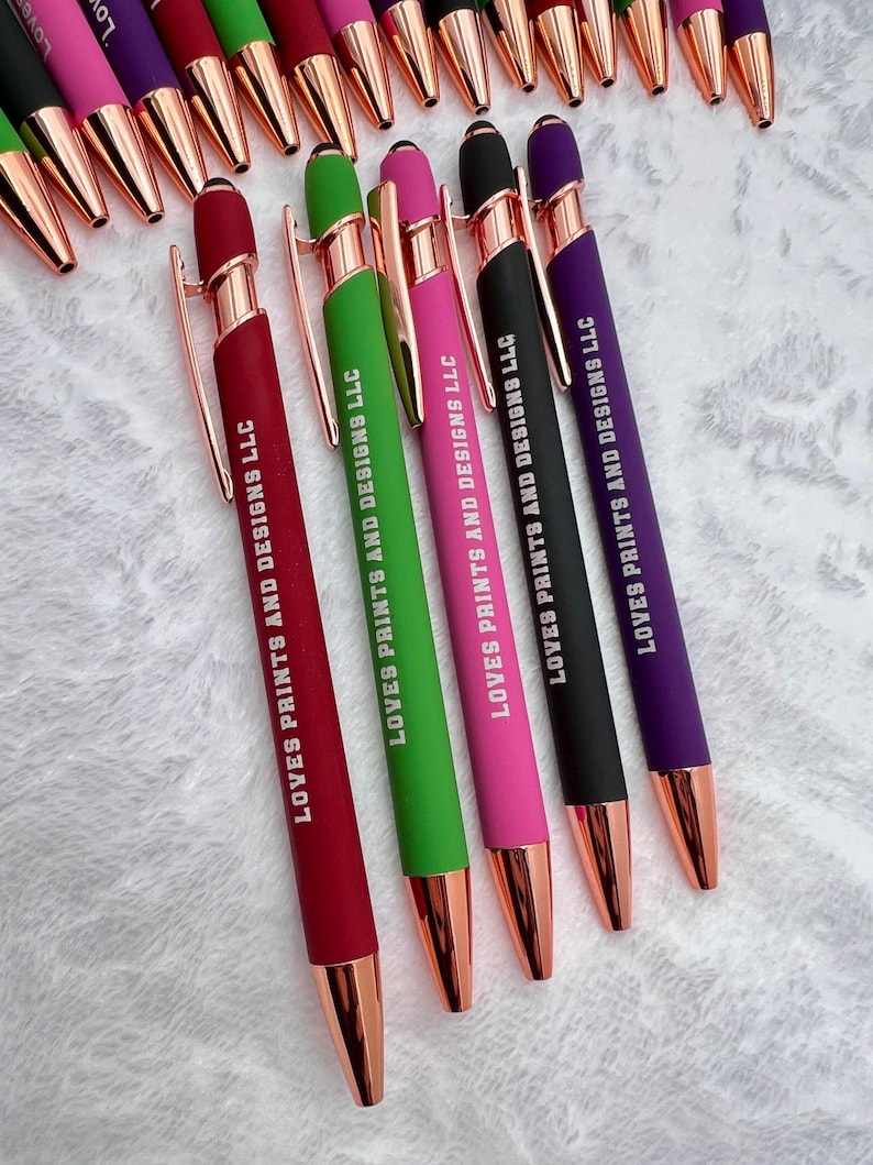 Pu&ograve; includere: Primo piano di sei penne con dettagli in oro rosa. Le penne sono rosse, verdi, rosa, nere e viola. Ogni penna ha il testo "LOVES PRINTS AND DESIGNS LLC" stampato sul lato.