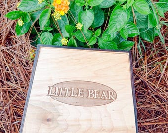 May include: A square wooden box with a light brown finish and a dark brown border. The lid features an oval engraving in the center with the text LITTLE BEAR in a stylized font. The box is positioned against a background of green foliage.