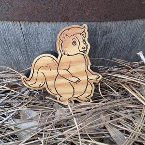 May include: A flat, light brown wooden cutout shaped like a cartoon squirrel. The squirrel is depicted in profile with a large, bushy tail, small ears, and a detailed outline burned into the wood grain, showing a standing posture with front paws.