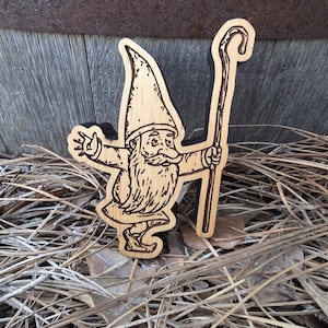 May include: A flat, laser-cut wooden gnome figure featuring a dark-edged silhouette. The gnome wears a tall, pointed hat, has a long beard, and holds a tall walking staff in one hand while extending the other arm outward in a waving gesture.