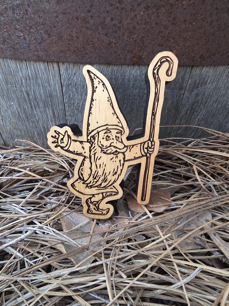 May include: A flat, laser-cut wooden gnome figure featuring a dark-edged silhouette. The gnome wears a tall, pointed hat, has a long beard, and holds a tall walking staff in one hand while extending the other arm outward in a waving gesture.