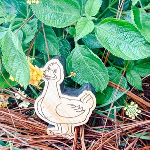 May include: A light-colored wooden cutout shaped like a duck with dark engraved outlines defining its features, including a beak, eye, wing, and feet. The duck figure has a smooth, natural wood grain texture and is positioned upright.
