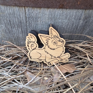 May include: A light-colored wooden cutout featuring a laser-engraved illustration of a long-haired dog with large pointed ears and a curled tail, lying down while holding a small stick between its front paws, with a black backing layer.