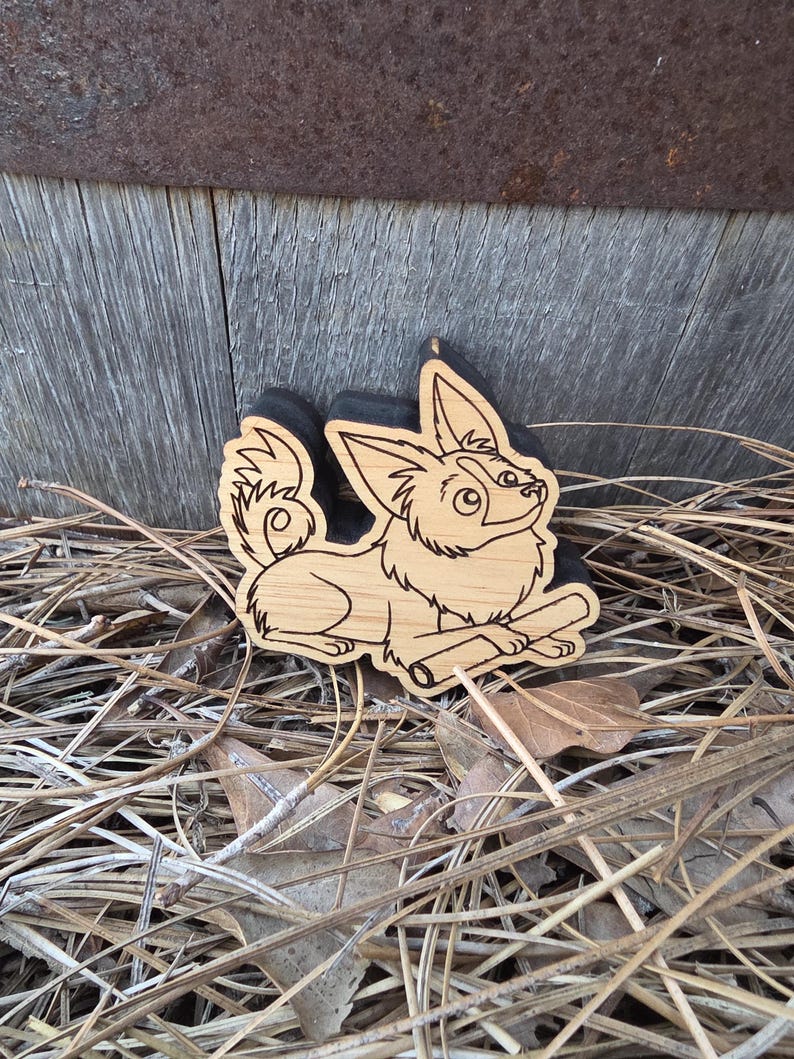 May include: A light-colored wooden cutout featuring a laser-engraved illustration of a long-haired dog with large pointed ears and a curled tail, lying down while holding a small stick between its front paws, with a black backing layer.