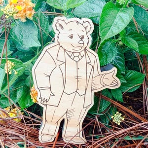 May include: A light-colored wooden cutout featuring a laser-engraved illustration of a bear standing upright. The bear is depicted wearing a formal three-piece suit, including a jacket, vest, and tie, with one arm extended in a gesturing motion.