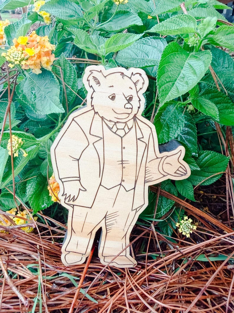May include: A light-colored wooden cutout featuring a laser-engraved illustration of a bear standing upright. The bear is depicted wearing a formal three-piece suit, including a jacket, vest, and tie, with one arm extended in a gesturing motion.