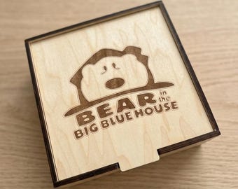 May include: A square wooden box with a sliding lid featuring a laser-engraved design of a bear peeking out from behind a house shape. The text "BEAR in the BIG BLUE HOUSE" is engraved below the bear illustration in a bold, sans-serif font.