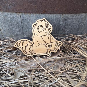 May include: A light-colored wooden cutout shaped like a cartoon raccoon with dark engraved outlines. The raccoon is depicted in a seated position with its striped tail curled behind it, featuring detailed facial features and fur texture lines.