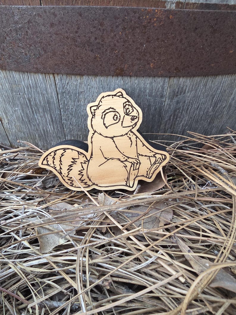 May include: A light-colored wooden cutout shaped like a cartoon raccoon with dark engraved outlines. The raccoon is depicted in a seated position with its striped tail curled behind it, featuring detailed facial features and fur texture lines.