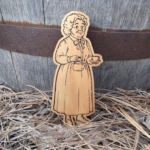 May include: A laser-cut wooden cutout featuring a detailed illustration of an older person with curly hair, wearing a long-sleeved dress with a bow at the collar, holding a small teapot in one hand and a teacup in the other, with dark etched lines.