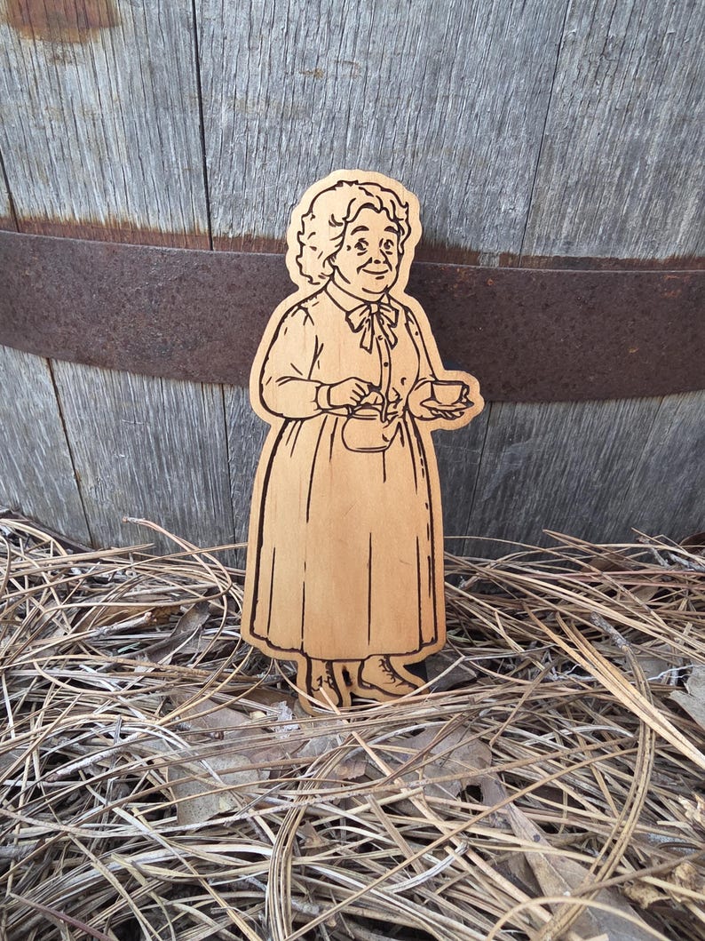 May include: A laser-cut wooden cutout featuring a detailed illustration of an older person with curly hair, wearing a long-sleeved dress with a bow at the collar, holding a small teapot in one hand and a teacup in the other, with dark etched lines.