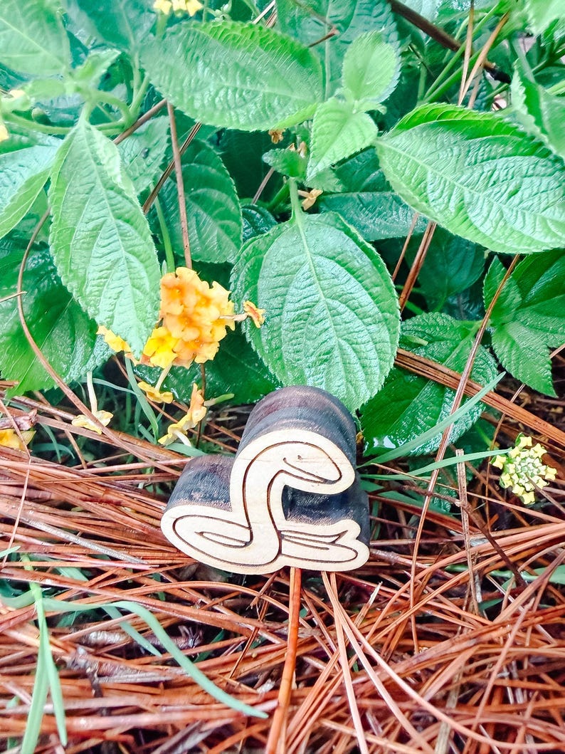 May include: A dark brown wooden stamp featuring a carved snake design on its face. The stamp has a cylindrical handle and a flat base with the snake outline clearly etched into the wood, showing a coiled body and a raised head profile.