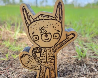 May include: A light-colored wooden cutout featuring a laser-engraved illustration of a cartoon fox-like creature with large eyes, pointed ears, and a bushy tail. The character is standing upright with one arm raised in a wave, displaying detailed line work.