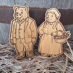 May include: Two light brown wooden cutouts featuring laser-engraved illustrations of anthropomorphic bears. One bear wears a suit, vest, and glasses, while the other bear wears a bonnet and dress, holding a basket filled with flowers and produce.