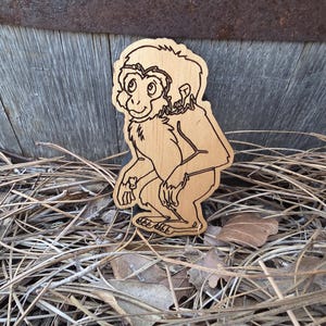 May include: A light-colored wooden cutout shaped like a monkey, featuring dark laser-engraved outlines that define the facial features, fur texture, and body posture of the primate. The figure is depicted in a crouching position with a gentle expression.